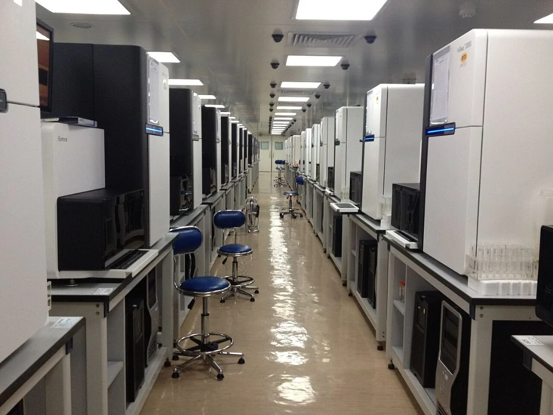 A long corridor lined on both sides with white sequencing machines and lab stools.