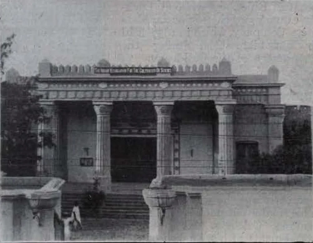 A low colonnaded building behind a wall and gate, with steps up to its entrance.