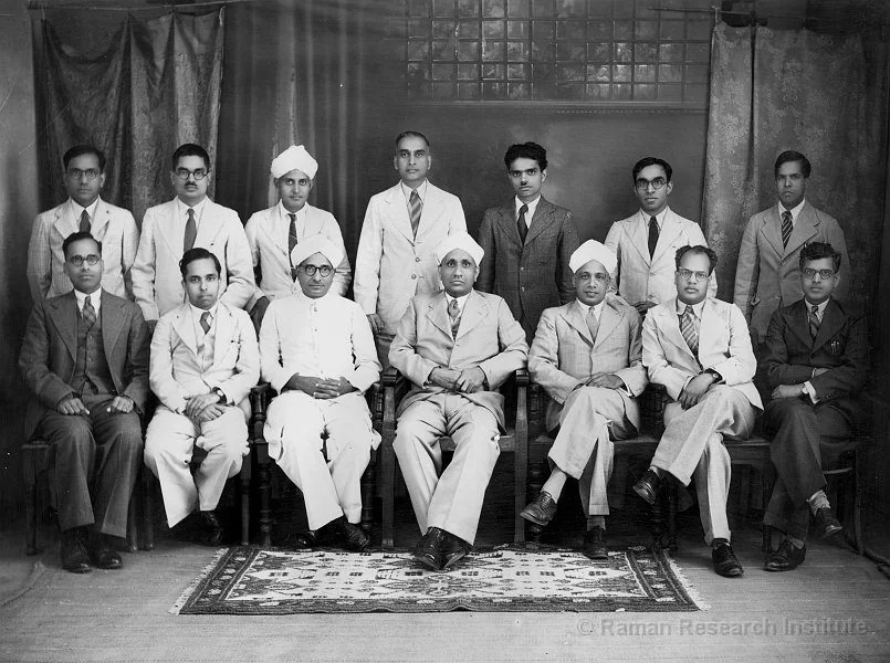Group photograph of fourteen men, seven seated and seven standing, on a patterned rug in front of a curtain.