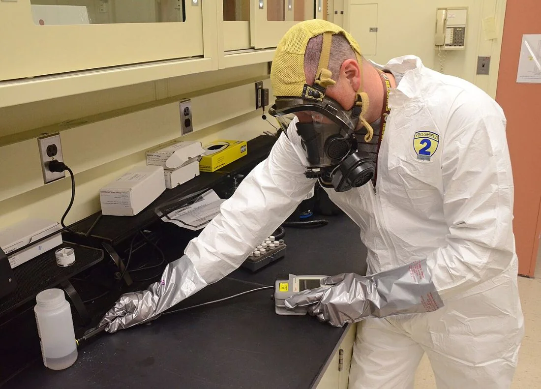 A man in a protective suit and respirator holds a handheld instrument against a sample bottle on a lab bench.