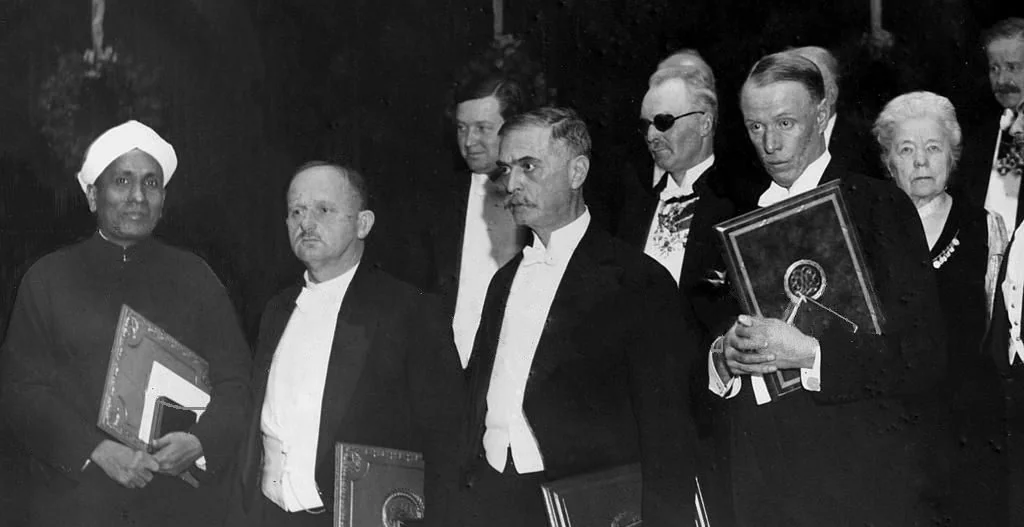 Four laureates holding Nobel diplomas, with others behind them. Raman, in a turban and closed coat, stands at the left.